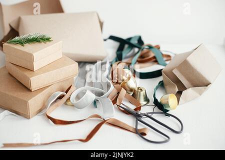 Verpackung stilvolle Öko weihnachtsgeschenke Komposition. Einfache Bastelschachteln, Samtbänder, weihnachtsglocken, Tannenzweig und Scheren auf weißem Backgroun Stockfoto