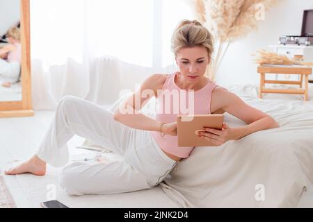 Eine schöne schlanke Frau sitzt am Morgen auf dem Boden, schauen Sie sich das digitale Tablet an Stockfoto