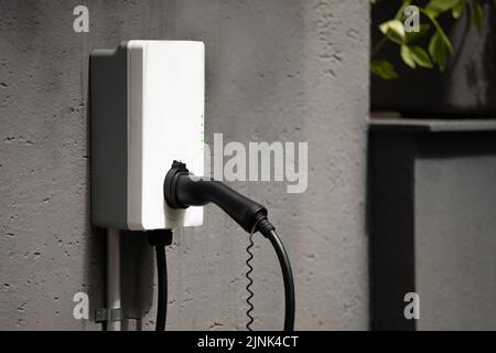 Ein Steckerhalter für die Wandmontage Ladestation mit Kabel an der Wanddose, die an der Betonhauswand montiert ist Stockfoto
