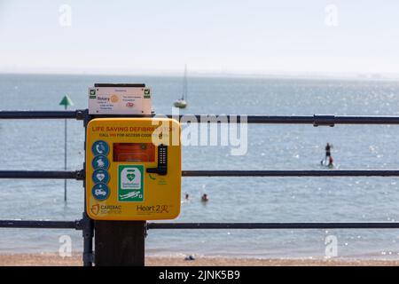 Ein öffentlicher lebensrettender Defibrillator in einem verschlossenen Kasten auf Metallgeländern mit Strand- und Meereshintergrund Stockfoto