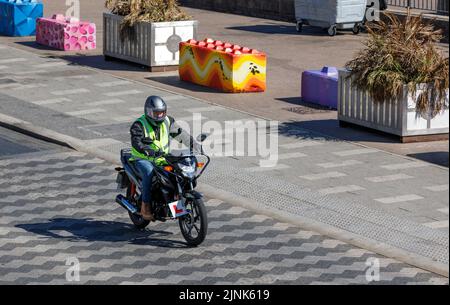 Ein Motorradfahrer, der mit Schutzkleidung und reflektierender Jacke auf einem Honda CB125F unterwegs ist, fährt auf einer Straße mit kariertem Pflaster Stockfoto