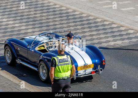 Ein Sicherheitsbeamter schaut auf ein blaues AC Cobra Auto mit weißem Streifen, das an einem heißen, sonnigen Tag entlang einer englischen Straße fährt, hohe Aussicht von oben Stockfoto