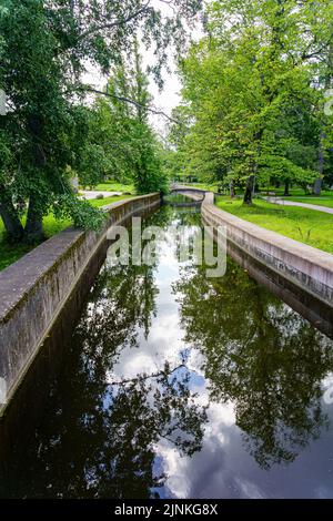 Kleine Brücke über ruhigen Wasserkanal im Park neben großen Bäumen. Stockfoto