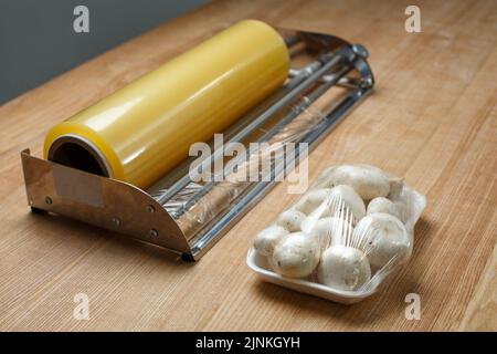 Nahaufnahme von Champignons auf Kunststofftablett in transparenter Frischhaltefolie verpackt. Frisch verpackte Pilze auf dem Tisch in der Nähe einer großen Rolle Lebensmittelverpackungsfolie. Co Stockfoto