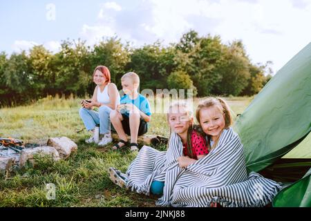 Zwei kleine umarmende Mädchen Schwestern auf dem grünen Gras neben dem Zelteingang des Lagers, fröhlich lächelnd. Schwester mit Bruder, der hinten auf dem Holzbuch sitzt und trinkt Stockfoto