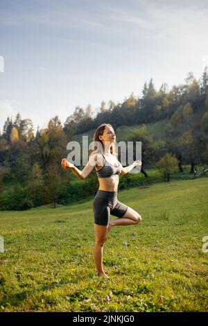 Eine junge Frau mit geschlossenen Augen meditiert, macht Atemübungen und hält das Gleichgewicht auf einem Bein. Stockfoto