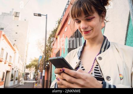 Junge lateinische kaukasische Transgender-Frau aus Argentinien, die auf der Straße steht und Nachrichten auf ihrem Telefon sendet, auf den Bildschirm schaut und mit einem lächelt Stockfoto