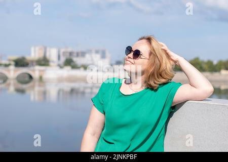 Porträt eines Mädchens in einer Sonnenbrille, eine grüne Bluse, die am Flussufer steht und ihr Haar mit der Hand hält. Ein Mädchen geht durch die Straßen des Cit Stockfoto