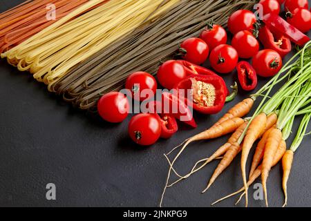 Hausgemachte bunte Pasta mit natürlichen Zutaten auf einem schwarzen Betontisch. Nudeln mit Tomaten, Karotten, Paprika. Vorbereitung zum Kochen zu Hause Stockfoto