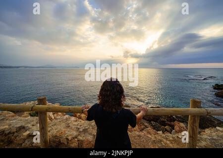 Frau, die von einem Aussichtspunkt aus auf den Horizont blickt, um den goldenen Sonnenuntergang über dem Meer zu genießen. Stockfoto
