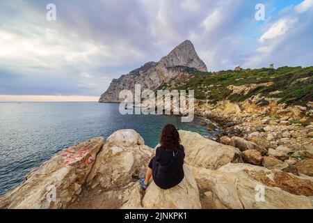 Frau mit umgedrehten Rücken und sitzend auf einigen Felsen, den Sonnenaufgang über dem Horizont des Meeres zu beobachten Stockfoto