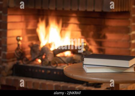 Bücher auf einem kleinen Tisch vor einem Kamin mit Kamin im Winter gestapelt. Stockfoto