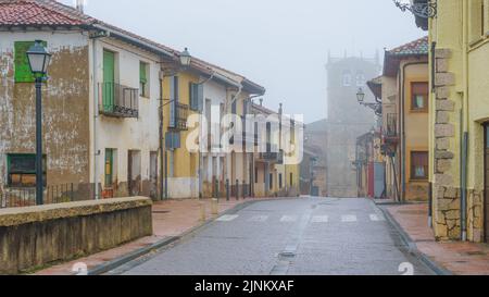 Gasse aus alten Häusern mit einer Kirche im Hintergrund an einem Tag mit dichtem Nebel und mystischer Atmosphäre. Riaza Segovia. Stockfoto