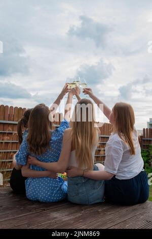 Rückansicht einer Gruppe junger Frauen mit langen Haaren, die klirrende Gläser mit Weißwein-Champagner auf dem Hintergrund des Himmels aufragen. Stockfoto