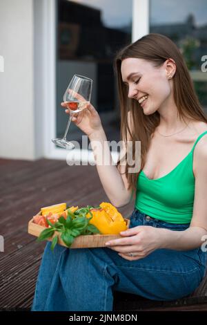 Porträt einer jungen lächelnden schönen Frau, die auf einer Holzverande sitzt und ein Glas Weißwein-geschnittenes Gemüse hält. Stockfoto
