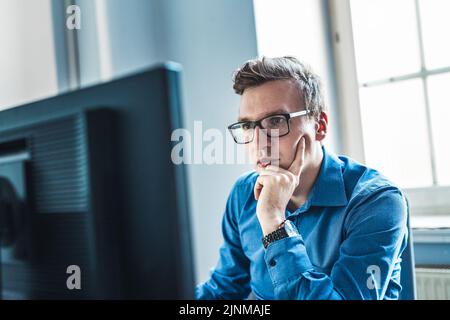 Hübscher junger Geschäftsmann mit Brille sitzt an seinem Tisch im Büro und schaut sich den Bericht auf seinem Computerbildschirm an. Stockfoto
