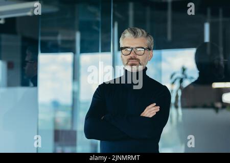 Porträt eines ernsthaften und fokussierten Geschäftsmannes mit gekreuzten Armen, eines Mannes, der im Büro arbeitet und eine Brille trägt und die Kamera anschaut, eines erfolgreichen, nachdenklichen Investors Stockfoto