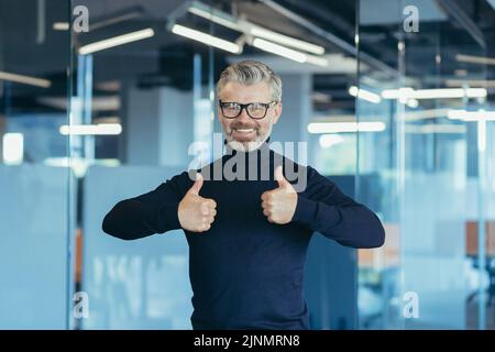 Porträt eines erfolgreichen Geschäftsmannes, eines älteren grauhaarigen Mannes im Büro, der zufrieden lächelt und die Kamera anschaut, wobei der Investor mit dem Daumen nach oben zeigt Stockfoto