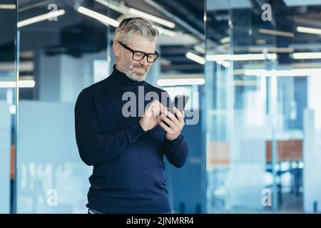 Leitender grauhaariger Geschäftsmann im Büro mit Telefon, glücklicher Investor, der die Nachricht liest und lächelt, Broker bei der Arbeit, der in einer Brille auf das Fenster schaut Stockfoto