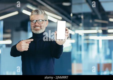 Leitender erfolgreicher grauhaariger Geschäftsmann im Büro, der der Kamera einen Telefonbildschirm mit weißem Hintergrund zeigt und mit dem Finger auf den Kopierbereich zeigt Stockfoto