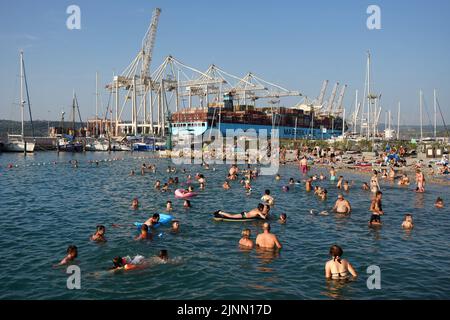 Menschen schwimmen an der felsigen Küste von Koper mit einem Frachtschiff im Hintergrund. Stockfoto