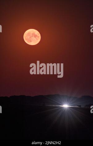 Bramble Hill, Bramshaw, Hampshire, Großbritannien, 12.. August 2022. Der Vollmond des Störs, der über der Landschaft des New Forest aufgeht, mit Autoscheinwerfern, die unten vorbeifahren. Kredit: Paul Biggins/Alamy Live Nachrichten Stockfoto