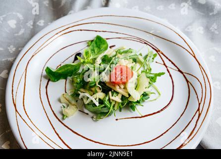 Gesunde Essenslöffel Salatsellerie und Apfelarugula mischen sich auf einem weißen flachen Teller, der mit Balsamico-Sauce auf einer grauen Stoffserviette auf dem hellen B dekoriert ist Stockfoto