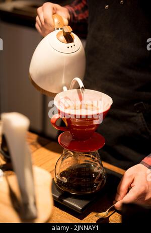 Der Barista schüttet heißes Wasser aus und macht Kaffee nach der Trichtermethode. Der Barista arbeitet hinter dem Tresen. Stockfoto