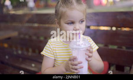 Das kleine Mädchen trinkt einen Milchshake durch einen Strohhalm. Nahaufnahme Porträt von niedlichen Mädchen auf Parkbank sitzen und trinken Milchshake. Odessa, Ukraine Stockfoto