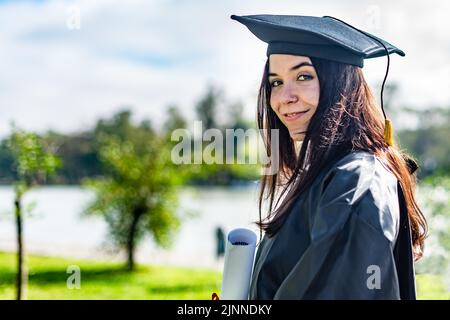 Happy Caucasian graduierte Mädchen mit langen braunen Haaren, mit einem Diplom und Blick auf die Kamera Stockfoto