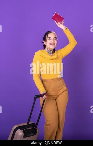 Junge glückliche Frau Reisende mit Koffer, während sie ihren Pass vor purpurem Hintergrund zeigt Stockfoto