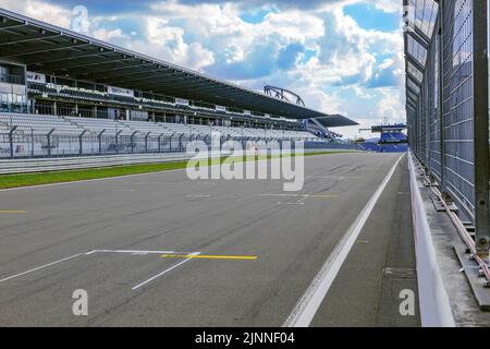 Blick durch den Sicherheitszaun von der Boxengasse auf die leere Start-Ziel-gerade Rasterspur mit Markierungen für die Startposition, Haupttribüne links Stockfoto