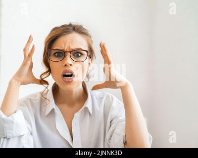 Nahaufnahme eines Porträts einer jungen verärgerten, wütenden Frau, die die Hände in wütender Geste hält. Junge Hündin mit roten Haaren in weißem Hemd. Negative menschliche Emotionen, f Stockfoto