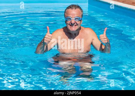 Ein weißer Mann mittleren Alters mit Daumen nach oben im Wasser eines Schwimmbades, mit einem sehr lustigen Ausdruck und einer Schwimmbrille Stockfoto