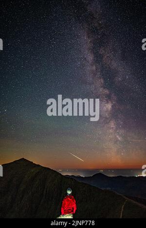 Mann (Sterngucker), der den klaren Sternenhimmel der Nacht, die Milchstraße und den Feuerball, den Sternschnuppen (Meteor über dem Snowdon-Gipfel im Snowdonia-Nationalpark) beobachtet Stockfoto