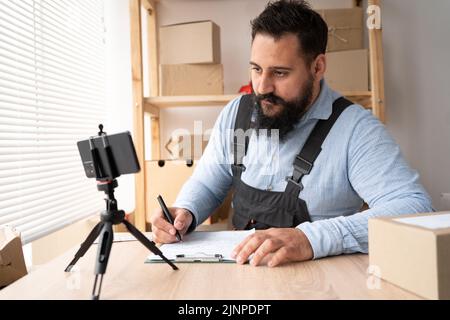 Ein junger Mann spricht mit dem Kunden über einen Videoanruf am Telefon, um den Warenauftrag zu überprüfen. Indische Geschäft männlich Paketboxen vorzubereiten, Überprüfung E-Commerce-Versand Stockfoto