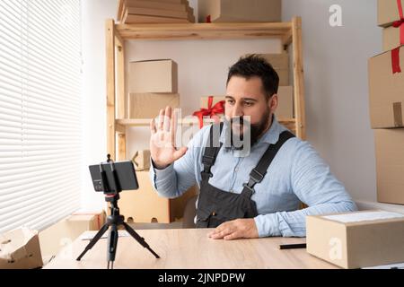 Junger indischer Geschäftsmann, der Smartphone im Lager verwendet, Berater des Online-Shops winkt Hallo an die Smartphone-Kamera. KMU, online einkaufen Stockfoto