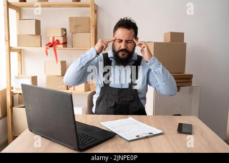 Junger indischer Mann, der am Laptop arbeitete, nachdem er das Paket zur Lieferung verpackt hatte. Inhaber von Kleinunternehmen, die im Büro arbeiten. Unternehmer mit Kopfschmerz denken Stockfoto