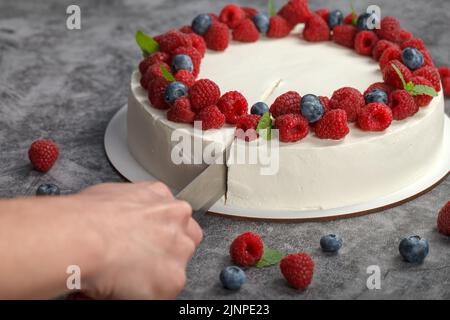 Der frisch gebackene Himbeerkuchen, der mit Himbeer-Beeren und Minzblüten verziert ist, wird mit einem Gebäckmesser geschnitten. Stockfoto