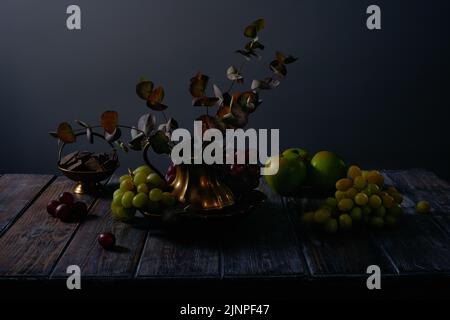 Fruchtstillleben mit Trauben, Äpfeln, Schokolade und antiken Kupfergeschirr auf dunklem Holztischhintergrund Stockfoto