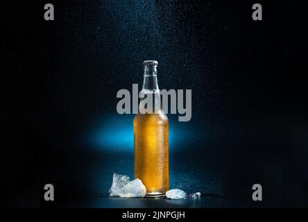 Bierflasche, Spritztropfen auf schwarzem und blauem Hintergrund. Eine Bierflasche mit spritzenden Tropfen mit Eiswürfeln auf dunklem Grund. Die Flasche Ka Stockfoto