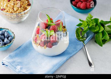 Gesundes Frühstück in einem Glas Parfait. Joghurt mit Müsli und Beeren auf einer blauen Serviette. Frische Himbeeren und Heidelbeeren mit hausgemachtem Müsli und y Stockfoto