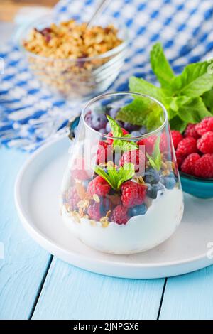 Gesunde Mahlzeit aus Müsli, Joghurt und Früchten auf bemalten Brettern. Diätdessert mit Joghurt, Müsli und Granatapfelfrüchten in einem Glas. Gesundes Frühstück Stockfoto