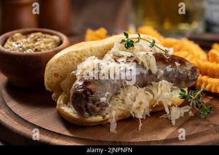 Nahaufnahme von Bratwurst und Sauerkraut auf einem Brötchen mit Senf und pommes auf einer Holzplatte Stockfoto