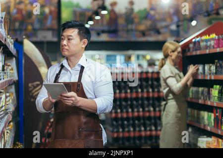 Zwei Arbeiter in einem Supermarkt in der Lebensmittelabteilung arrangieren Produkte, ein asiatischer Mann mit einem Tablet-Computer und eine Frau in Schürzen unter den Produkten in den Regalen und Regalen Stockfoto