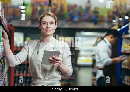 Porträt einer erfahrenen Verkäuferin in einem Supermarkt, die Managerin schaut auf die Kamera und lächelt, hält einen Tablet-Computer in den Händen, die Verkäuferin ist mit Waren im Laden in den Regalen Stockfoto