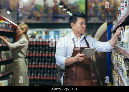 Zwei Arbeiter in einem Supermarkt in der Lebensmittelabteilung arrangieren Produkte, ein asiatischer Mann mit einem Tablet-Computer und eine Frau in Schürzen unter den Produkten in den Regalen und Regalen Stockfoto