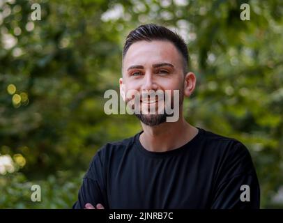 Porträt eines jungen, gutaussehenden Mannes mit Bart. Junger Mann im schwarzen Hemd lächelt und schaut in die Kamera. Outdoor Natur Hintergrund Stockfoto
