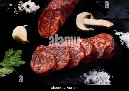 Iberischer Chorizostock in Scheiben geschnitten auf schwarzem Hintergrund mit Knoblauch, Salz, Petersilie und einer Scheibe Pilz Stockfoto