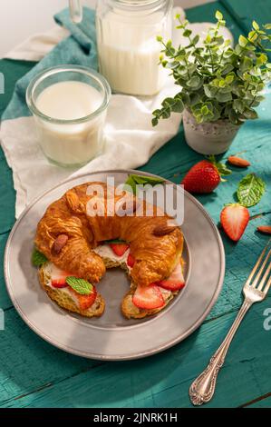 Wunderschön servierte Frühstückstisch für eine Person. Croissant mit frischen Erdbeeren, Mandeln, einem Glas Milch und Blumendekor im Innenbereich. Französische Küche Stockfoto